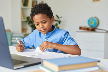 Curious black schoolboy with curly hair sitting at table and checking notes in workbook while doing...