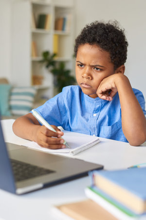 Frowning thoughtful black boy with curly hair sitting at table and listening to tutor during video...