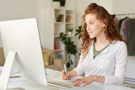 Smiling attractive redhead student girl sitting at table and making notes while enjoying online...