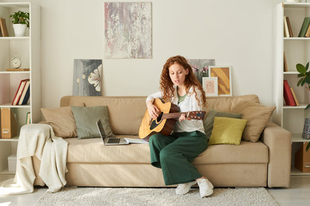 Focused redhead girl sitting in living room and watching video lesson while learning to play guitarの写真素材