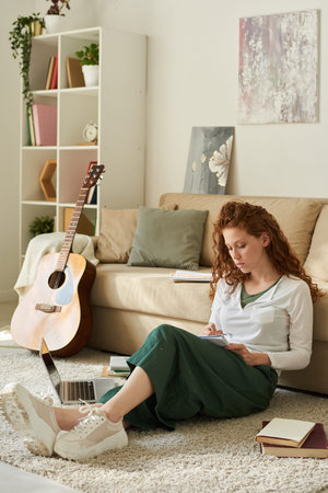 Concentrated redhead student girl with curly hair sitting on floor and doing home task in living roomの写真素材