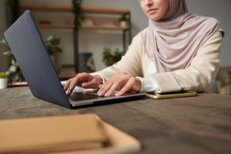 Modern Muslim woman wearing hijab sitting at office desk typing something on her laptop, copy spaceの写真素材