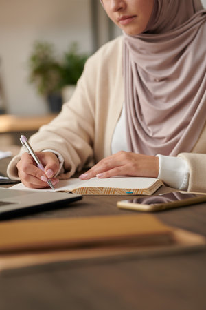 Middle Eastern woman wearing headscarf and abaya sitting at office table writing notes in her notebook, vertical head cropped shotの写真素材