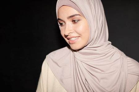Head and shoulders portrait of cheerful beautiful Muslim girl wearing hijab standing against black...