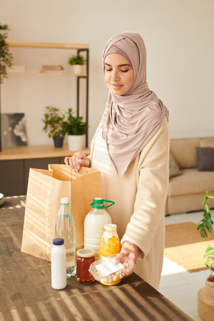 Vertical medium long shot of beautiful young Muslim woman wearing hijab standing at table taking out food delivered to herの写真素材