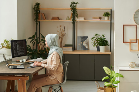 Young Middle Eastern professional programmer wearing light blue hijab sitting at desk at home...
