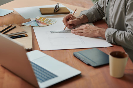 Close-up of unrecognizable man sitting at desk and using ruler while drawing apartment plan