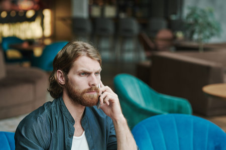 Displeased young bearded man sitting in modern cafe and waiting for answer while calling by cellphoneの写真素材