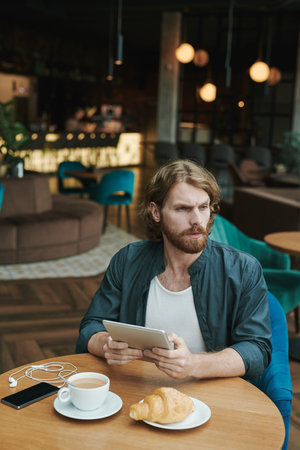 Serious handsome guy with beard sitting at table in cafe and drinking coffee while reading mailの写真素材