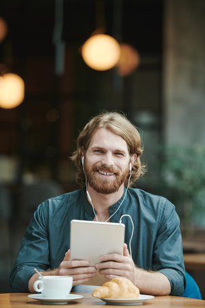 Confident young man in casual shirt sitting in armchair and looking away while talking by phone in...