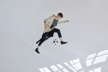 Caucasian tattooed man in sporty clothes kicking soccer ball in large room with white walls illuminated by sunlightの写真素材