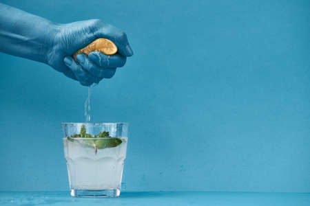 Preparing delicious mojito: unrecognizable man squeezing lemon juice into faceted glass with homemade beverage, blue backgroundの写真素材