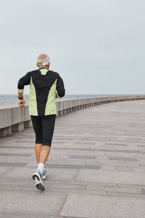 Unrecognizable active elderly man jogging along seaside in morning vertical shot