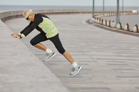 Horizontal side view shot of sporty senior man doing morning aerobic exercises outdoors
