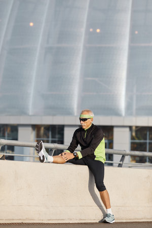 Vertical shot of slim elderly man doing ligaments stretching exercise outdoors