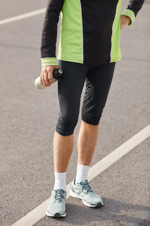 Lower body shot of unrecognizable fit man wearing tracksuit standing on track with bottle of water