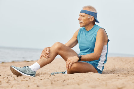 Horizontal shot of aged man wearing sporty outfit sitting on sand looking away