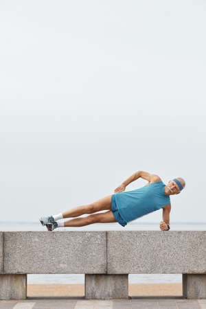 Vertical shot of strong senior man wearing sporty summer outfit doing side plank exerciseの写真素材
