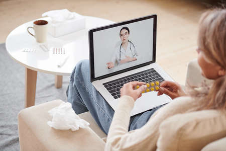 High angle shot of woman sitting on sofa staying at home talking to doctor on video call using laptopの写真素材
