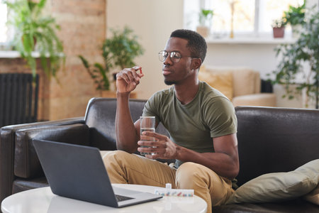 Young adult African American man wearing eyeglasses sitting on sofa in front of laptop holding glass...