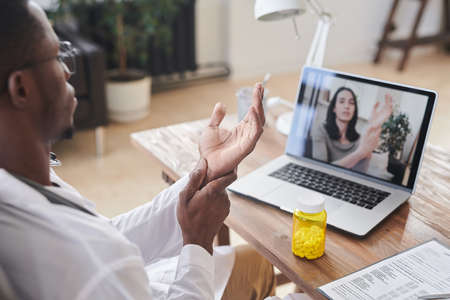 Rear view shot of modern African American doctor sitting at desk in front of laptop showing during online video callの写真素材