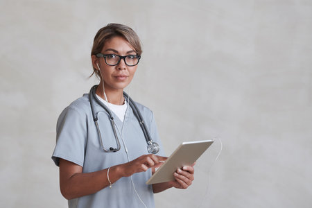Horizontal medium studio portrait of modern female doctor wearing eyeglasses and earphones using...