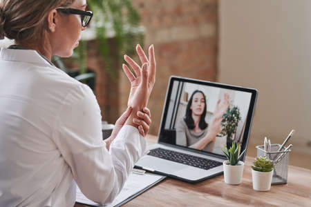 Modern medical worker wearing white coat sitting at desk in loft office having online appointment with patient using laptopの写真素材