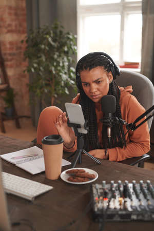 Vertical shot of modern young woman with afro braids wearing headphones sitting at desk in living room at home adjusting camera settings on smartphone to shoot video for blogの写真素材