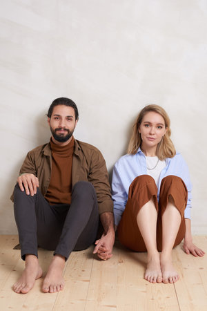 Portrait of young man and woman wearing casual clothes sitting on floor holding hands looking at camera, copy spaceの写真素材