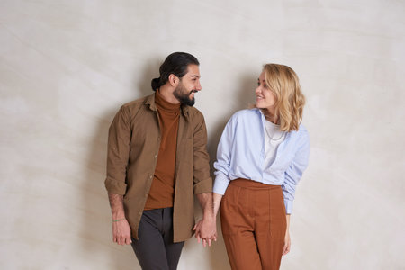 Medium long portrait of lovely young couple standing against wall holding hands and flirting, copy...