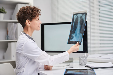 Horizontal side view medium portrait shot of mature Caucasian female doctor wearing white coat sitting at desk in her office looking at X-ray shotの写真素材