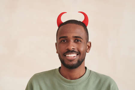 Portrait of cheerful young black bearded man with red devil horns standing against beige background, Halloween conceptの写真素材