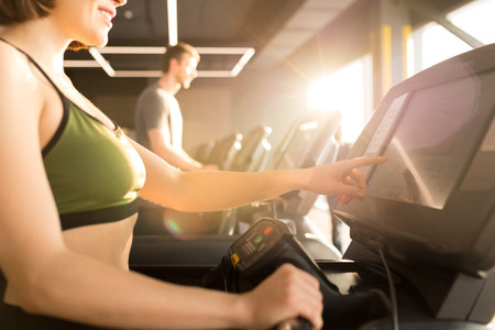 Young woman in sports bra choosing a program on treadmill for her cardio training with man running...