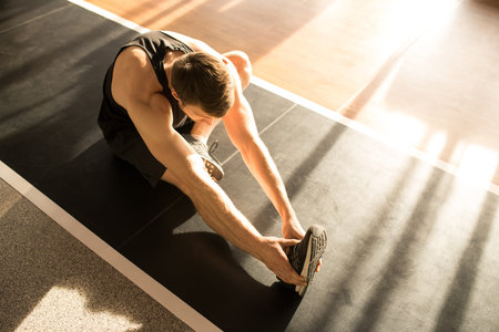 Fitness young man sitting on the floor on exercise mat and doing stretching exercises in gym