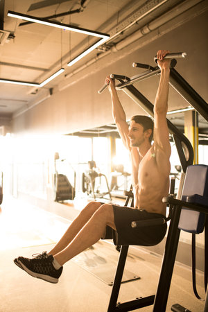 Handsome shirtless athlete doing pull ups exercises on sports equipment in sport club