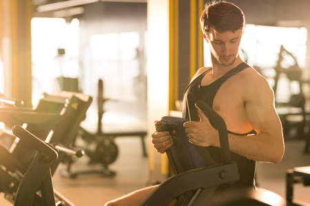 Young man training on sports equipment and looking at his muscles during strength training in health clubの写真素材