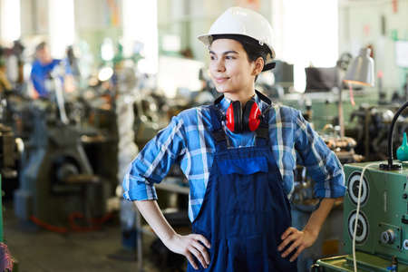 Content purposeful skilled female factory worker with ear protectors on neck wearing hardhat and overall holding hands on hips and looking away proudlyの写真素材