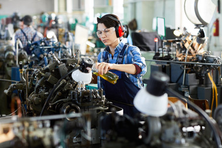 Serious concentrated lady engineer in ear protectors and protective goggles pouring lubricant for...