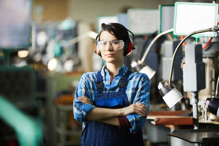 Serious gritty woman in safety goggles and ear protectors standing at industrial shop and crossing arms on chest while looking at cameraの写真素材