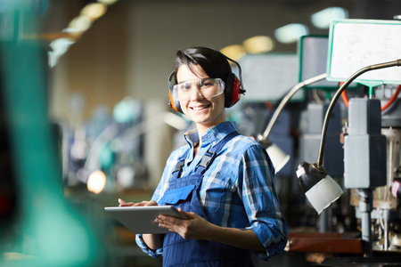 Smiling excited young female technician in protective goggles and ear protectors standing in factory...