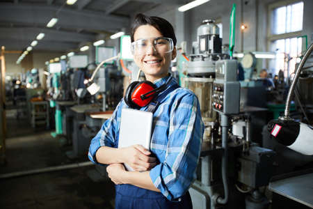 Cheerful confident masculine lady worker of watch factory wearing ear protectors on neck and safety goggles standing against automatic machines and embracing tabletの写真素材