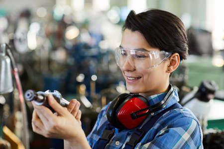 Smiling satisfied young woman in transparent goggles wearing ear protectors examining metal drill bit while choosing detail for metalworkingの写真素材