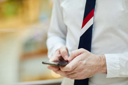 Close-up of unrecognizable bank employee in necktie and white shirt standing and making note in smartphoneの写真素材