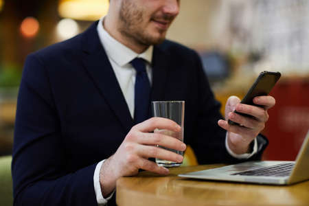 Close-up of content businessman in suit sitting at table and drinking water while reading Internet news on smartphoneの写真素材