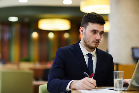 Serious concentrated handsome business expert in suit sitting at table in restaurant and editing information in papers while using laptopの写真素材