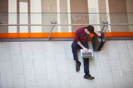 Top view of serious young man in trousers and casual shirt sitting on floor and leaning on carry-on luggage in airport hallの写真素材
