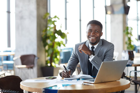 Content successful young African-American businessman in suit sitting at table in restaurant and analyzing papers while preparing reportの写真素材