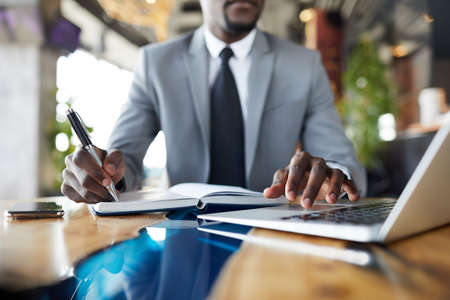 Close-up of black businessman in suit sitting at table in cafe and examining data on laptop and making notes in organizer, he preparing business planの写真素材