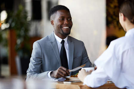 Smiling excited young African-American businessman sitting at table and eating salad while discussing project over business lunchの写真素材