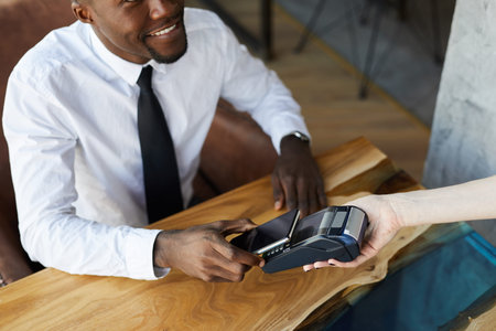 Close-up of smiling young black businessman in white shirt and necktie sitting at table paying for...